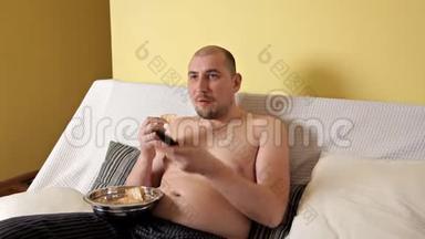 懒洋洋的，秃头的，胖乎乎的，没有外套的胖子正在看电视，在沙发上喝着薯片的可口可乐。 胖子